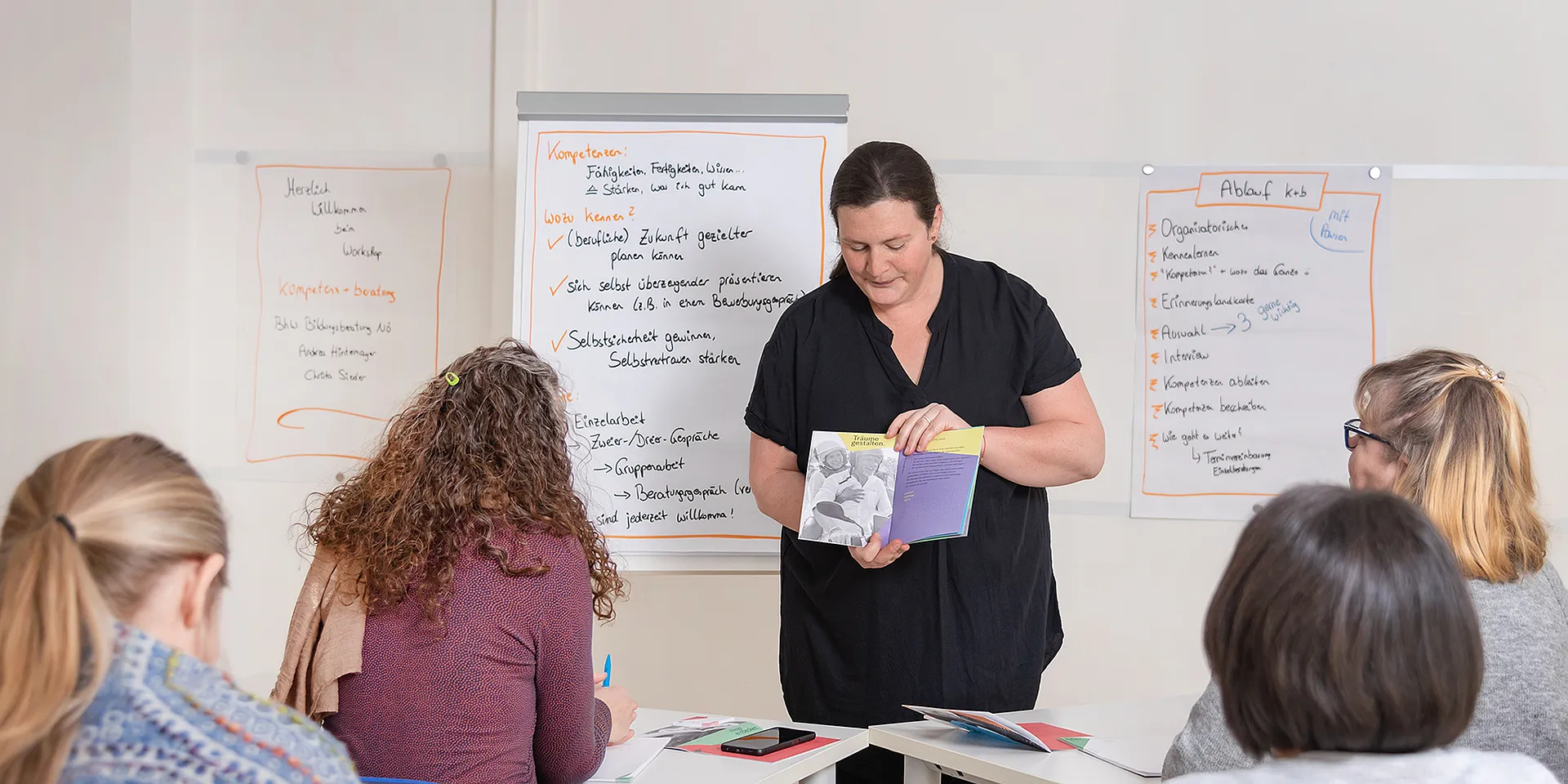 Bildungsberaterin Christa Sieder zeigt einen Folder interessierten Zuhörerinnen und Zuhörern, im Hintergrund steht ein Flipchart mit Informationen.