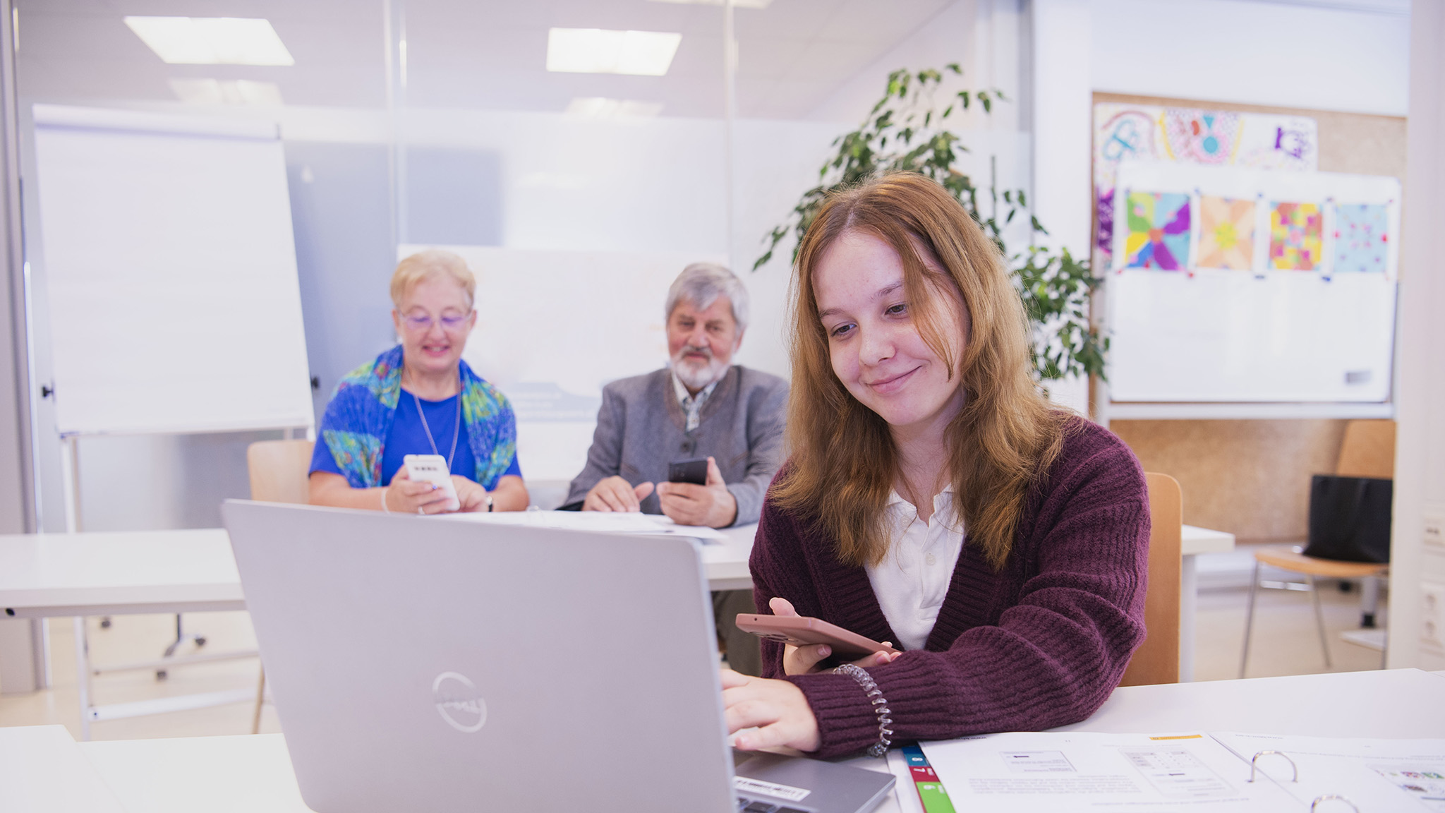 Eine junge Frau sitzt vor einem Laptop und hält ein Smartphone in der Hand, während sie konzentriert arbeitet und lächelt. Hinter ihr sitzen zwei ältere Personen, die ebenfalls mit ihren Smartphones beschäftigt sind, in einem hellen Schulungsraum.